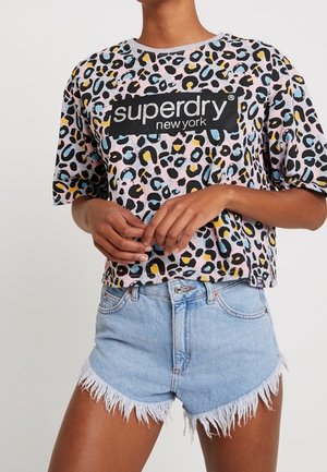 Person in einem Superdry New York Cropped Top mit Leopardenmuster und hellblauen, ausgefransten Denimshorts, stehend mit gefalteten Händen.