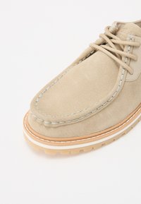 Chaussure en suédine beige avec un bout arrondi, un détail de couture bleue et une semelle en caoutchouc brun clair présentant un motif de grip texturé.