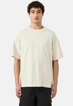 Hombre con cabello rizado, vistiendo una camiseta beige holgada con el logotipo "Patrick" y pantalones cortos cargo negros, de pie contra un fondo liso.