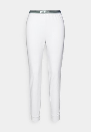 Balti medvilniniai sportiniai kelnių su žaliais ir tamsiai mėlynais dryžiais ant juosmens, kuriuose įspaustas "LACOSTE" logotipas. Prigludę šlaunys su raukiniais.