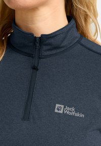 Nahaufnahme einer Frau, die einen dunkelgrauen Jack Wolfskin Fleece mit Viertelreißverschluss trägt, wobei das Markenlogo auf der Brust sichtbar ist.