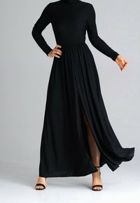 Zwarte maxi-jurk met lange mouwen, hoge halslijn, elastische tailleband en een zij-split. Geccombineerd met zwarte sandalen met hak. Glad, vloeiend materiaal.