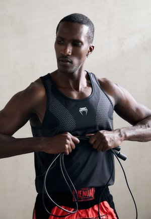 Schwarzes sportliches Tanktop mit Mesh-Einsätzen und einem Logo, getragen von einer Person, die ein Springseil hält; rote Shorts mit einem Markengummizug.