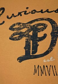 Camiseta de algodón naranja con gran texto negro desgastado, con detalles de "D" y "MMVII". Incluye costuras de acento blancas.