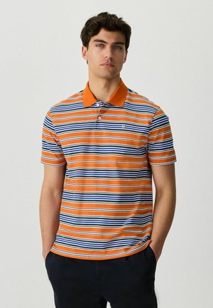 Junger Mann mit einem horizontal gestreiften Polohemd in Orange, Blau, Grün und Weiß, dazu schwarze Hosen, steht mit den Händen in den Taschen.