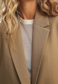 Béžové sako s výstřihem se zářezy, hladká látka, vrstvené přes lehký texturovaný svetr. Zdůraznění čistých linií a přizpůsobeného střihu.