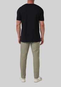 Man staat met zijn rug naar de camera, gekleed in een zwart T-shirt met korte mouwen, lichtgroene broek en witte sneakers, op een effen grijze achtergrond.