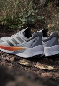 Baskets de sport grises avec des accents orange. Elles présentent un dessus texturé, trois rayures gris foncé et un design de semelle distinct et courbé.