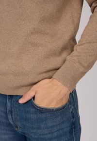 Pull en tricot beige avec des poignets côtelés, associé à un jean bleu foncé. La main est posée dans la poche du jean, affichant un style décontracté.