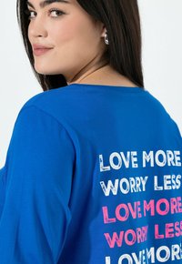 Blaues Baumwoll-T-Shirt mit dem Text "LIEBE MEHR, SORGE WENIGER" in Weiß und Pink, lockere Passform mit weitem Rundhalsausschnitt und überschnittenen Schultern.