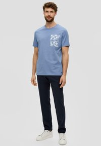 Hellblaues Baumwoll-T-Shirt mit weißen Blumenmuster auf der Tasche, kombiniert mit dunklen Hosen und weißen Turnschuhen. Figurbetontes Design, kurze Ärmel.