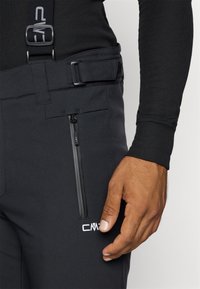 Pantaloni neri impermeabili con bretelle regolabili, dotati di una tasca laterale con zip e un logo sul lato anteriore inferiore. Materiale liscio e resistente.