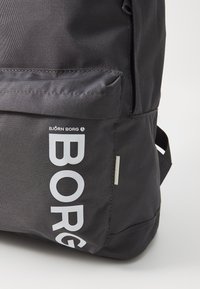 Sac à dos en tissu noir avec une grande poche zippée sur le côté, texture lisse et lettrage blanc "BORG" audacieux sur la surface avant.