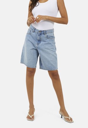 Kobieta ubrana w jasnoniebieskie bermudy jeansowe, biały top na ramiączkach oraz białe sandały na obcasie, stojąca z nogami rozstawionymi na białym tle.