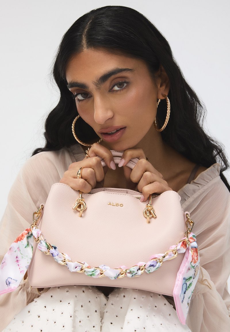 Femme aux longs cheveux foncés et grandes boucles d’oreilles en anneau tenant un sac à main ALDO rose clair avec un foulard à motif floral sur les anses en chaîne dorée.