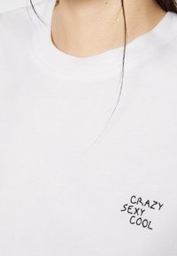 Bílá tričko s kulatým výstřihem a černým vyšitým textem "CRAZY SEXY COOL" na levé části hrudníku, s pramenem tmavých vlasů viditelným poblíž krku.
