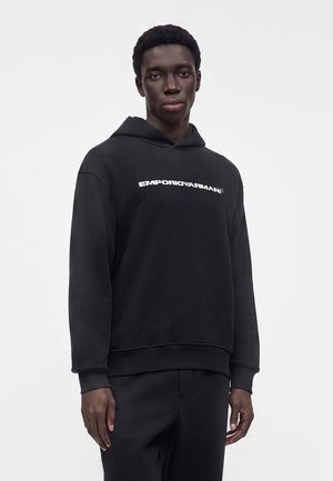 Junger Mann trägt schwarzen Hoodie mit weißem "EMPORIO ARMANI"-Text und steht vor einem einfarbigen hellgrauen Hintergrund.