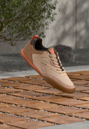 Baskets Calvin Klein beige et marron avec lacets sur des dalles en bois à l’extérieur près d’un mur en béton et d’un arbuste vert.