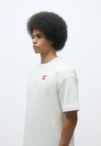 Light cream short-sleeve t-shirt featuring a small red square logo on the left chest, made from soft material with a relaxed fit.
