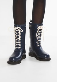 Bottes de pluie en caoutchouc navy avec une finition brillante, lacets blancs, œillets en métal et semelle noire. Présente un logo en haut de l'arbre.