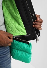 Ręce rozpinające czarną kurtkę, ukazujące zieloną pikowaną podszewkę oraz przymocowaną zieloną ocieplaną warstwę wewnętrzną na szarym tle.