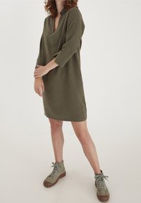 Robe vert olive, longueur genou, avec un décolleté en V et des manches trois-quarts. Tissu doux avec une coupe décontractée, assortie à des bottes vertes à lacets.