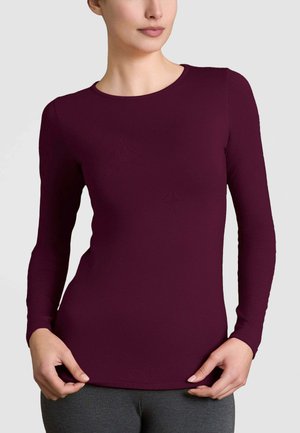 Camicia a maniche lunghe bordeaux con scollatura rotonda, silhouette aderente e tessuto morbido; indossata su un pantalone grigio.