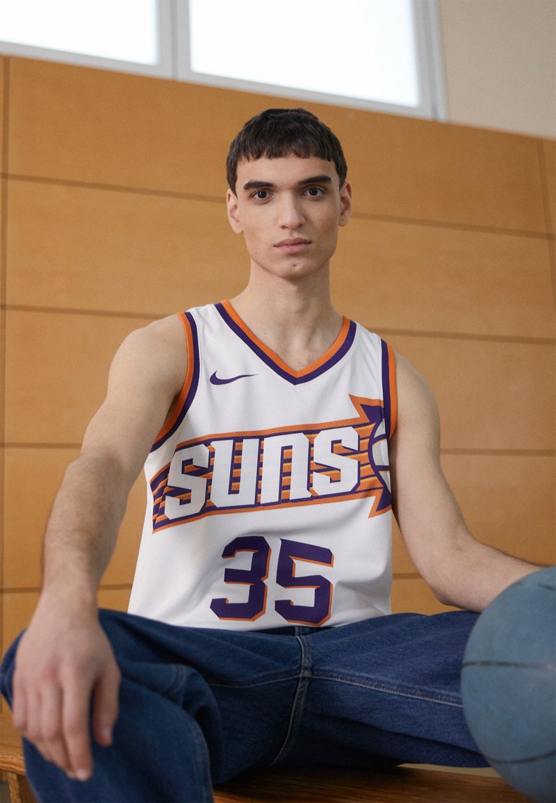 Maillot de basket-ball blanc avec des accents orange et violet, comportant le texte "Suns" et le numéro "35". Le modèle porte un jean bleu et tient un ballon de basket.
