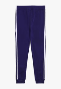 Lila träningsbyxor med vita sidostrick, elastisk midja, tapered ben design och mjuk textur. Inga ytterligare detaljer synliga.