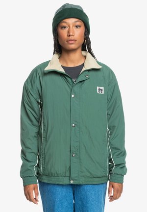 Green lightweight jacket with a beige collar, snap buttons, and white stitching accents; worn over a black shirt and blue jeans.