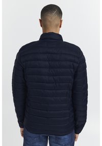 Marineblå pufferjakke med horisontalt quiltet mønster, stående krage og slim fit. Materialet ser glatt og lett ut.