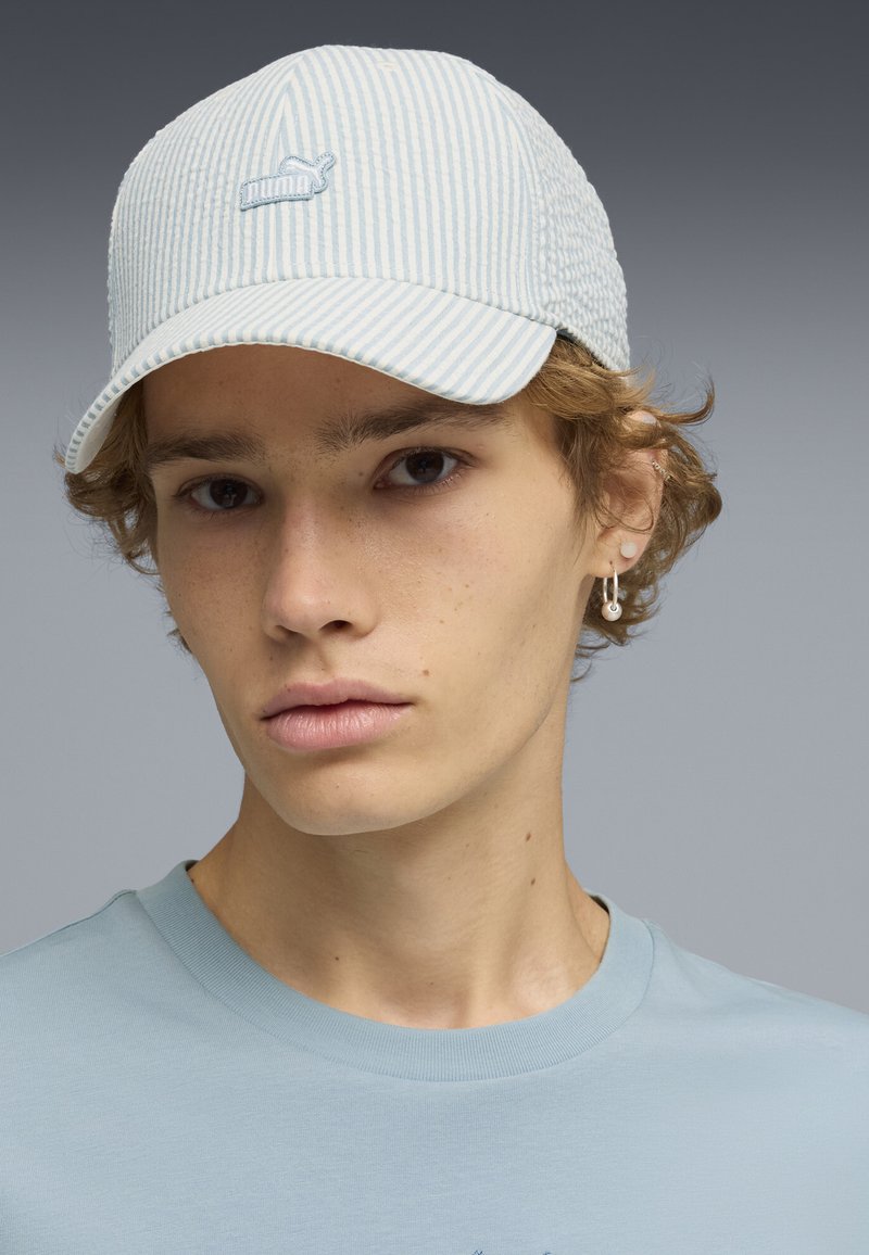 Jeune personne aux cheveux bouclés châtain clair portant une casquette rayée bleu clair, des boucles d'oreilles argentées en créole et un t-shirt bleu pâle à col rond.