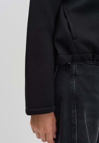 Zwarte sweatshirt met een omgeslagen mouw en zijzak, gecombineerd met high-waisted donkerdenim jeans, die een strakke en minimalistische stijl tonen.