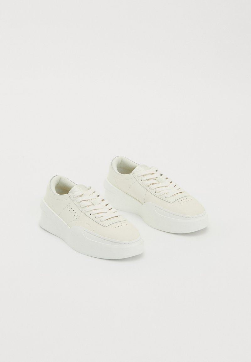 Hvide lavtoppede sneakers med tykke såler og snørebånd, der har perforeret detaljering på siderne og tåområdet, præsenteret på en hvid baggrund.