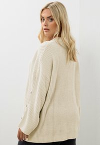 Sweter w kolorze kremowym, wykonany na drutach, o luźnym kroju, z teksturowanym wzorem i guzikami na rękawach. Miękka, średniej wagi tkanina.