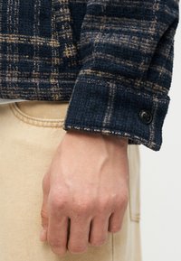 Κοντινό πλάνο ενός χεριού που φοράει μπεζ παντελόνι και μανίκι από μάλλινο σκούρο μπλε καρό σακάκι με μαύρο κουμπί στη μανσέτα.