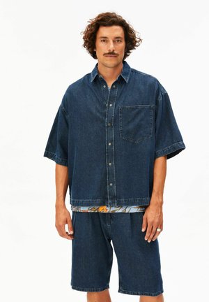 Mann mit lockigem Haar und Schnurrbart, der ein dunkelblaues übergroßes Jeanshemd und passende Shorts trägt und vor weißem Hintergrund steht.