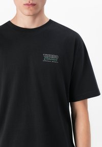 Sort bomulds T-shirt med korte ærmer. Forsiden har et lille logo i hvid og grøn tekst: "TURBO Classics EUROPEAN IMPORTS."