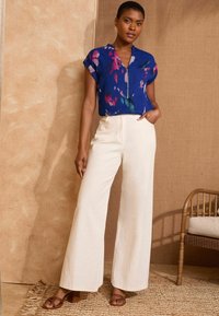 Blusa floreale blu a maniche corte, con zip frontale, abbinata a pantaloni bianchi a gamba larga. Il modello indossa sandali marroni con cinturini. Sfondo testurizzato.