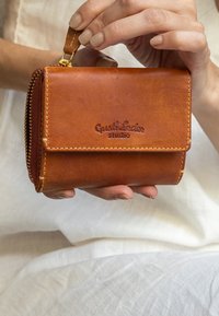 Bruine leren portefeuille met een gouden rits en een gegraveerd logo. Compact, rechthoekig ontwerp met een gladde textuur. Gestikte accenten zichtbaar.