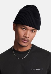 Căciulă tricotată ribbed, de culoare neagră, cu o potrivire strânsă. Purtați-o cu o cămașă închisă la culoare, ce are un logo mic alb și un lanț de argint.