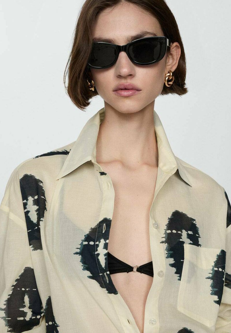 Durchsichtige beigefarbene Bluse mit schwarzen abstrakten Mustern und einer Brusttasche, kombiniert mit schwarzen Sonnenbrillen und einem schwarzen Bikinioberteil.