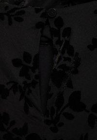 Tissu noir avec un motif floral en velours, doté d'un bouton et d'une petite ouverture fendue au centre.