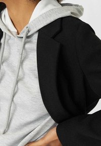 Blazer noir au design structuré porté par-dessus un sweat à capuche gris. Présente un cordon de serrage, une texture en coton et des détails de coutures visibles.