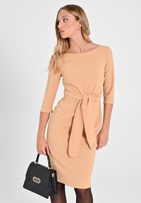 Beiges, tailliertes Kleid mit drei Viertelärmeln und einer vorderen Bindung. Kombiniert mit einer strukturierten schwarzen Handtasche mit einem goldenen Verschluss.