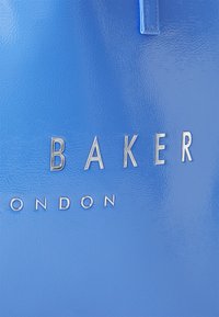 Modrá lesklá kabelka so strieborným nápisom "BAKER LONDON." Má hladkú textúru a štruktúrovaný dizajn.