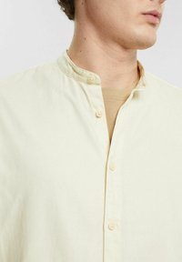 Camicia gialla chiara con colletto alla mandarina, design monopetto e bottoni coordinati. Tessuto morbido, vestibilità casual, indossata sopra una camicia beige.