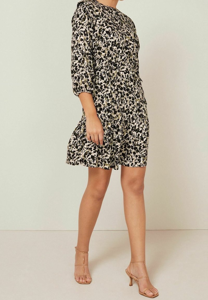 Femme portant une robe noire et blanche à motifs, longueur genou, manches trois-quarts, et des sandales à talons hauts beiges avec brides.