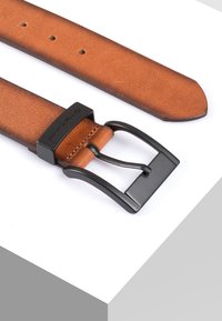 Ceinture en cuir tan avec une texture lisse et une boucle en métal noir. Elle dispose de plusieurs trous pour les ajustements et d'une extrémité effilée.
