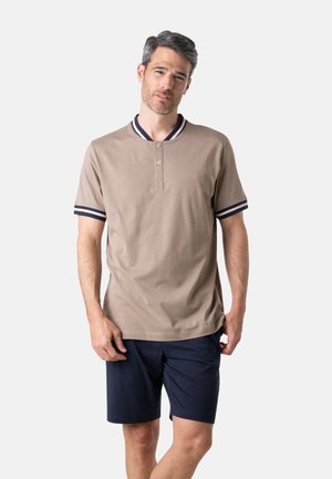 Homme portant un polo beige à manches courtes avec un col et des poignets rayés bleu marine, assorti à un short bleu marine arrivant aux genoux, debout devant un fond blanc.